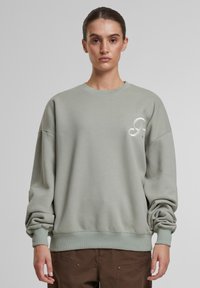 Lys grøn genser med rund hals, dropped shoulders, ribbede mansjetter og nederkant, med en subtil brodert logo på brystet.