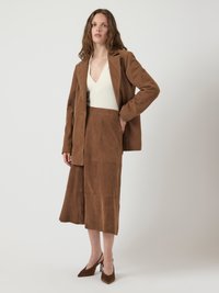 Mulher com cabelo solto e ondulado veste um blazer de camurça marrom e uma saia midi sobre uma blusa branca de decote em V, combinada com sapatos de salto alto pontudos marrons.