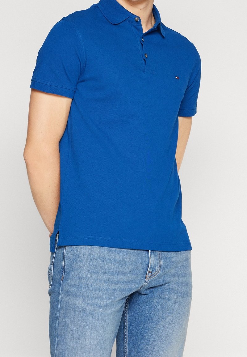 Homme portant un polo bleu à manches courtes et un jean bleu clair, les mains derrière le dos, debout devant un fond uni.