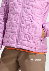 Vadderad rosa jacka med vadderad yta, frontdragkedja och elastiska ärmslut. Kombination med bruna byxor som har orange midjeband och vit logotyp.
