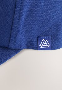 Tissu bleu à tissage serré avec des coutures visibles montrant une petite étiquette cousue avec un logo de montagne blanc sur fond bleu.
