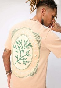 T-shirt beige avec un graphique botanique vert et du texte. Manches courtes, coupe décontractée, texture douce et encolure arrondie.