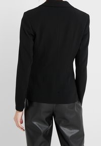 Zwarte op maat gemaakte blazer met een lange, getailleerde silhouet, notch revers en naaideetails. G gedragen over hoge zwarte nepleren broeken.