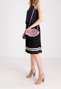 Violetin vaunge, joka on muotoiltu huuliksi, jossa on ketjusankka, käytettävänä mustan pleated-mekon ja beige perforoitujen korkokenkien kanssa.