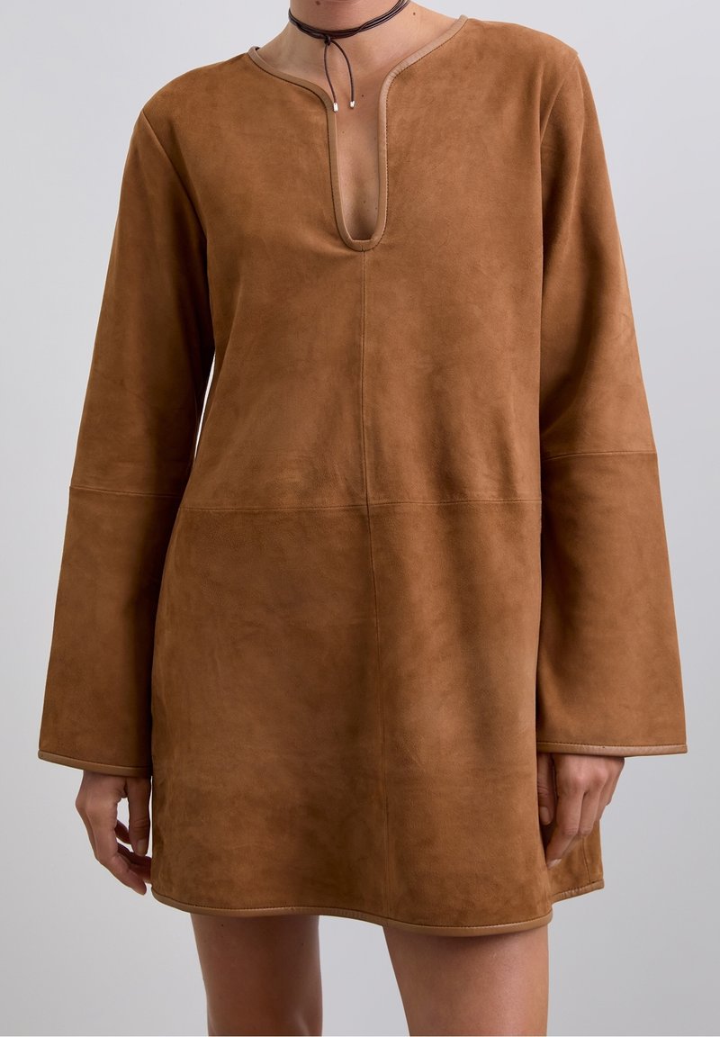 Robe longue en suède marron à manches longues avec une fente en V profonde portée par une personne, associée à un collier ras-du-cou fin noir avec de petites perles.