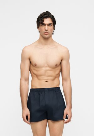 Homme torse nu aux cheveux courts et foncés portant un short noir à taille élastique, debout face à l'avant sur un fond clair uni.