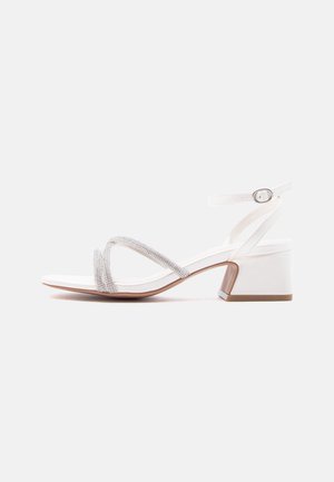 Hvid sandal med lav hæl, tynd ankelrem og to dekorative glitrende stropper foran mod en ensfarvet baggrund.