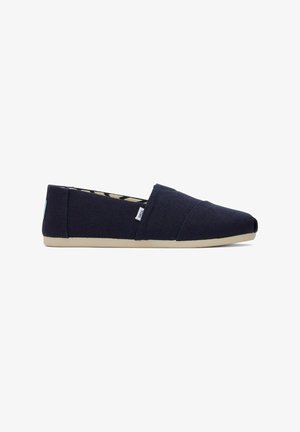 Navy blue slip-on canvas shoe with beige sole, featuring side stitch detailing and a small fabric brand tag.