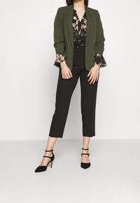 Olivgrön blazer över en svart blommig blus med volangärmar, i kombination med svarta cropped byxor och svarta klackskor med ankelsnören.