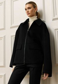 Sort lynlåsfælde jakke med stor krave, to frontlommer og brede ærmer. Båret over en lys turtleneck og parret med sorte bukser.