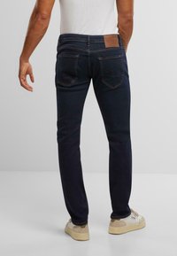 Tumši zila denima bikses ar taisnu piegriezumu, ar ādas zīmogu jostasvietā, klasiskām aizmugurējām kabatām un kontrastējošiem šuvumiem.