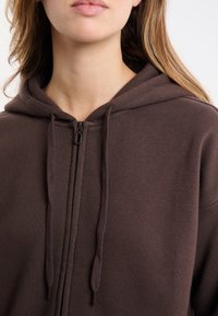 Brązowa bluza z kapturem na zamek, wykonana z miękkiego materiału, z kapturem, zamkiem z przodu i sznurkami przy kapturze. Prosty design bez wzorów.