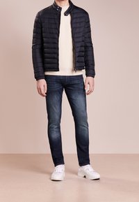 Pánská péřová bunda tmavě námořnické modré barvy, krémový svetr s žebrováním, tmavě modré džíny a bílé tenisky. Bunda má stojatý límec a zapínání na zip.