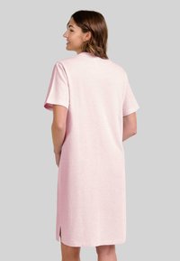 Mujer con cabello castaño hasta los hombros que lleva un vestido suelto de color rosa claro hasta la rodilla, con mangas cortas, de pie frente a un fondo gris liso.