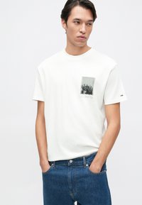 Camiseta blanca de algodón con un estampado rectangular en blanco y negro de un paisaje urbano etiquetado como "Nueva York" en cursiva, cuello redondo y mangas cortas.