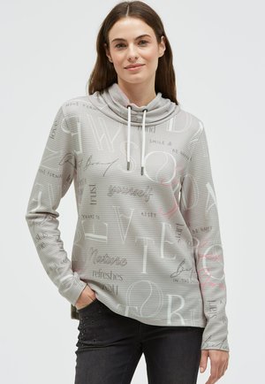 Young woman wearing a light gray striped hoodie with motivational words, paired with black jeans, standing against a plain background.