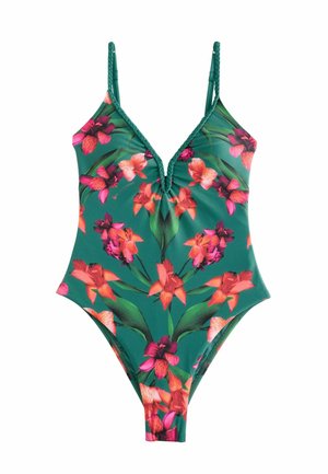 Femeie cu părul blond până la umeri, purtând un costum de baie verde întreg, cu modele florale roșii și roz, lângă un perete de piatră.