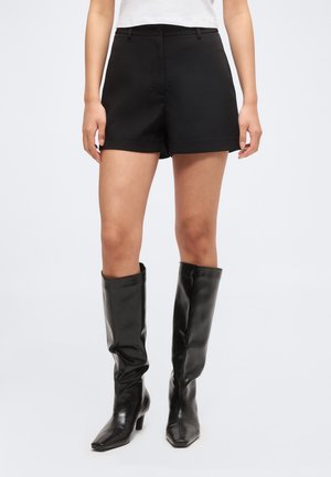 Femme portant un short noir sur mesure et des bottes en cuir noir hautes jusqu'aux genoux, debout devant un fond clair et uni.