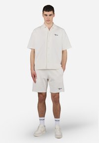 Camicia a maniche corte di un leggero color crema con motivo a rombi e ricami neri, abbinata a pantaloni corti coordinati e sneakers bianche.