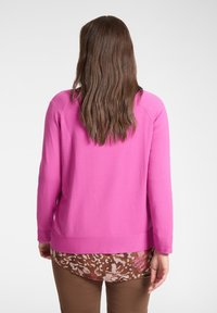 Różowy sweter z długimi rękawami i prostym dołem. Noszony na brązową bluzkę w wzory, z kwiatowymi motywami w jaśniejszych kolorach.