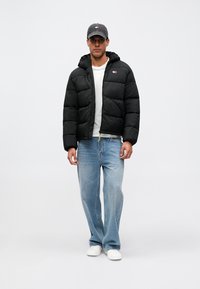 Bărbat îmbrăcat cu o geacă puffer neagră, cămașă albă, blugi deschiși la culoare, adidași albi și o șapcă gri, stând în fața unui fundal simplu.