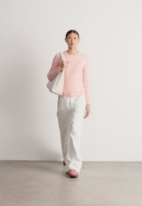 Svetloružový sveter s dlhými rukávmi a pleteným vzorom, kombinovaný s bielymi širokými nohavicami. Modelka nesie bielu tašku a nosí ružové topánky.