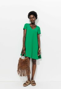 Robe verte à manches courtes en V, au design fluid. Accessoire : sac en suède marron avec des franges et des clous. Chaussures : sandales à glissière en imprimé léopard.