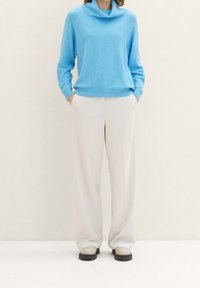 Ljusblå cowl-neck tröja i mjukt stickat tyg, tillsammans med krämfärgade byxor med vid ben. Platta skor med tjock sula i neutrala färger fulländar outfiten.