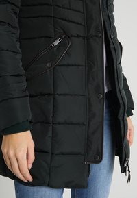 Veste noire matelassée avec poche latérale zippée et boutons-pression, portée avec un jean bleu, une main reposant à côté.