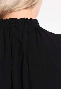 Black garment with a high, gathered neckline featuring a button closure at the back. The fabric appears smooth with a soft texture.