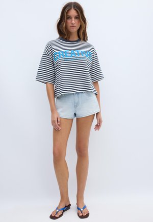 Jeune femme portant un t-shirt rayé noir et blanc "CREATIVE", un short en jean clair et des tongs bleues, debout devant un fond uni.