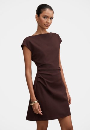 Mujer que lleva un vestido ajustado sin mangas de color marrón oscuro con ligeros pliegues en la cintura, complementado con pendientes y pulseras doradas.