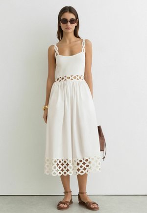 Kobieta w białej sukience bez rękawów z okrągłymi wycięciami na ramiączkach, talii i dole, brązowych sandałach, okularach przeciwsłonecznych i trzymająca brązową torebkę.