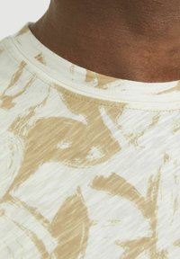Tela de patrón beige y crema con textura acanalada; escote redondo y material suave, mostrando diseños florales sutilmente mezclados con el fondo.