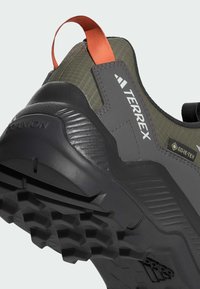 Zapatilla de senderismo GORE-TEX con parte superior sintética en gris oscuro, suela de goma texturizada y tira de agarre naranja; cuenta con un talón reforzado y un diseño elegante.