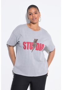 Camiseta de algodón gris con cuello redondo, que presenta un audaz texto rojo "STUDIO" y un gráfico de rayo con estampado de leopardo. Mangas cortas, corte relajado.