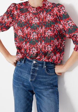 Frau trägt eine rote Bluse mit Blumenmuster und Puffärmeln, in hoch taillierte blaue Jeans gesteckt, Hände in den Hüften.
