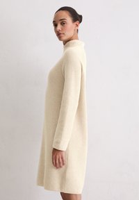 Sukienka z długim rękawem i wysoką szyją, wykonana z miękkiego kremowego dzianinowego materiału, o luźnym kroju i prążkowanej teksturze. Bezszwowa konstrukcja z bocznymi kieszeniami.