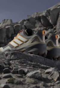 Παπούτσια για trail running με ανθεκτικές σόλες και μπεζ, γκρι και πορτοκαλί σχέδιο τοποθετημένα σε βραχώδες έδαφος κάτω από καθαρό ουρανό.
