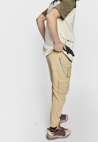 Persoon in beige cargobroek met kettingdetails, bruine en paarse sneakers, en een shirt met korte mouwen met een beige vest dat aan de onderkant van het vest trekt.