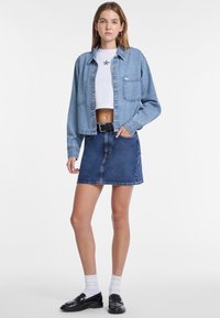 Giacca di jeans azzurro chiaro a metà vita con chiusura a bottoni e tasche, abbinata a una gonna di jeans blu scuro. Mocassini neri e calzini bianchi completano il look.