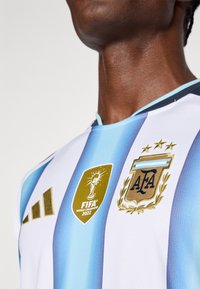 Balta un debeszilā svītrainā futbola krekls ar izšūtu zeltītu FIFA Čempioni 2022 logo un Argentīnas Futbola asociācijas emblēmu.