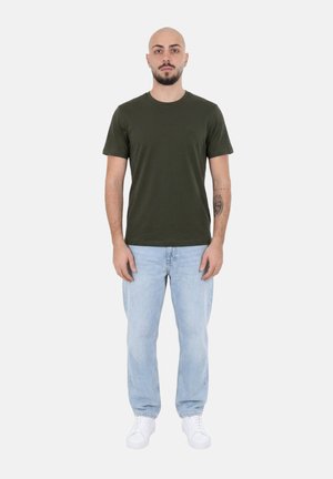 Uomo in piedi rivolto in avanti, indossa una t-shirt verde scuro, jeans azzurri, sneakers bianche, con un tatuaggio visibile sull’avambraccio sinistro.
