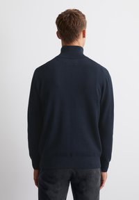 Marineblauwe gebreide trui met een hoge kraag, voorzien van een gestructureerd patroon en geribbelde manchetten, gecombineerd met donkere broek.