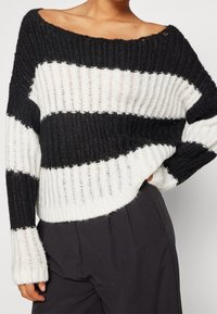 Pull en maille rayé noir et blanc à coupe ample, avec une texture côtelée, un col large et des manches longues légèrement évasées.