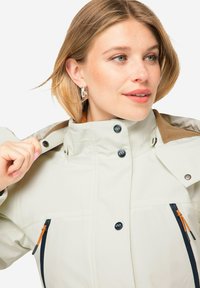 Vaalea kerma takki korkealla kauluksella, ruskea yksityiskohta kaulan lähellä, vetoketjulliset taskut ja mustat napit. Silkkinen pinta, jossa on vähäiset yksityiskohdat.