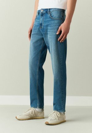 Mann trägt mittelblaue Jeans mit geradem Bein, weiße Socken und beige Sneakers, steht vor einer schlichten hellgrauen Wand.