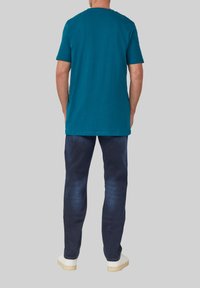 Türaqua Baumwoll-T-Shirt mit kurzen Ärmeln und Rundhalsausschnitt, kombiniert mit dunkelblauen Jeans und weißen Sneakers, von hinten betrachtet.