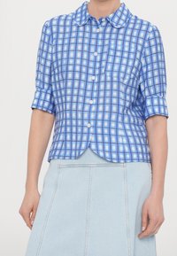 Camicia a quadri blu con colletto, maniche corte arrotolate, cinque bottoni e taschino sul petto sinistro. Realizzata in tessuto leggero.