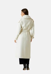 Lungo cappotto crema con collo in pelliccia, vita con cintura e dettagli ai polsi. Tessuto liscio, lunghezza al ginocchio, mostrato di spalle con stivaletti neri alla caviglia.